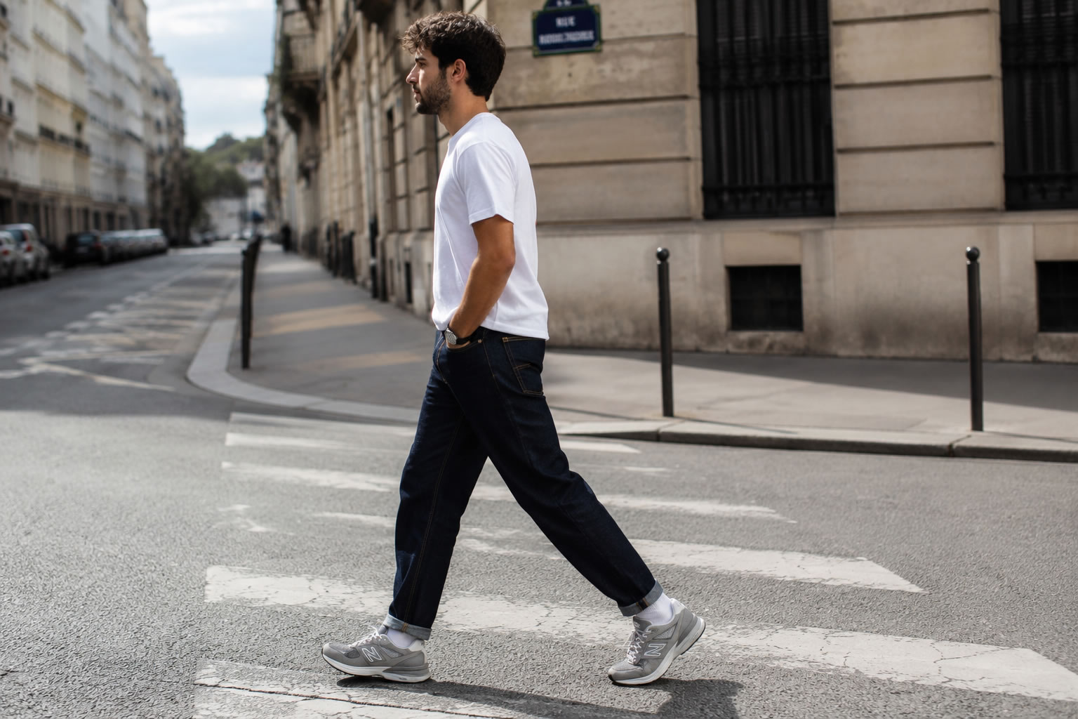 Homme portant des New Balance grises en ville | Look casual homme Homme marchant en ville avec un jean brut, un t-shirt blanc et des sneakers New Balance grises dans un style casual urbain.