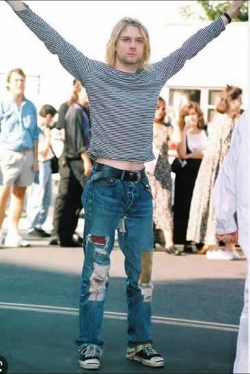Kurt Cobain avec des Converse One Star et un jean patchwork Kurt Cobain portant des Converse One Star, un jean patchwork et un t-shirt rayé lors d'une apparition publique.