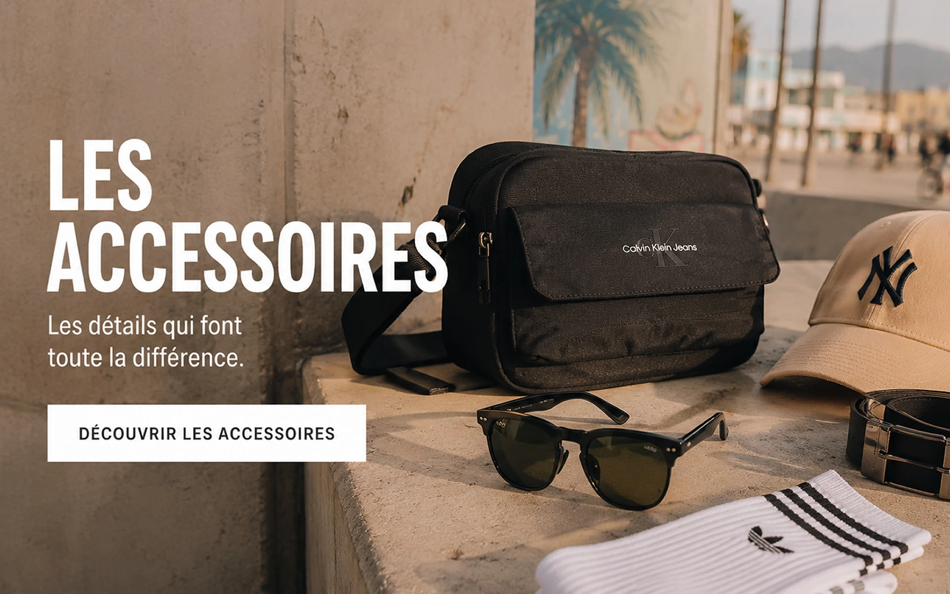 Accessoires Homme Tendance – Casquette, Sac et Lunettes Accessoires de mode pour homme avec sac bandoulière noir, casquette beige, lunettes de soleil et ceinture sur une terrasse ensoleillée.