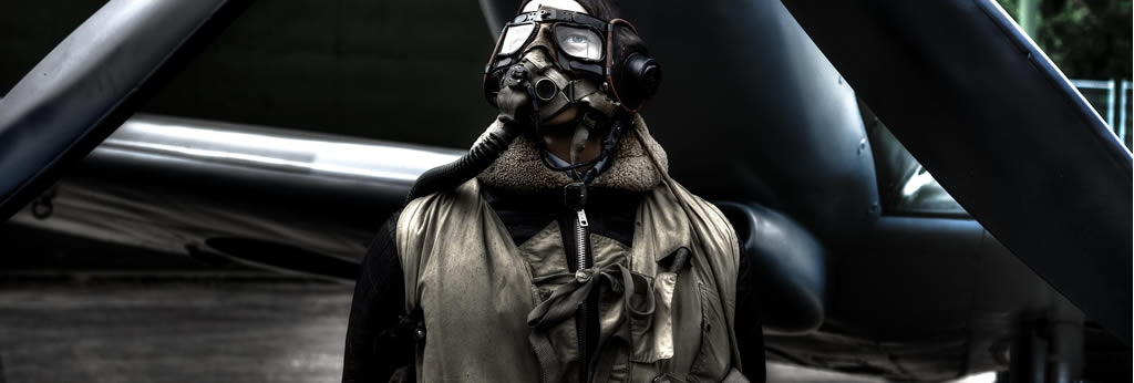Aviateur en tenue de vol ancienne avec casque, lunettes et masque devant l'hélice d'un avion.